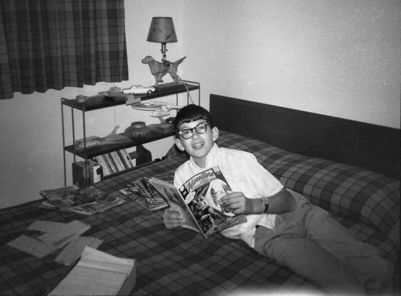 Young boy in glasses reads a copy of Adventure Comics in his bedroom ca. 1969